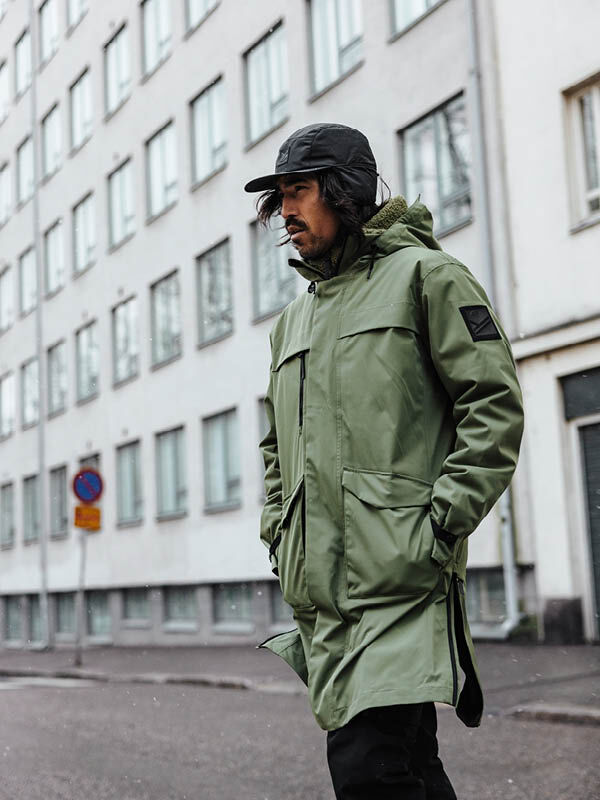 Person in a Halti Tokoi II DX Parka Jacket and black cap walks along a city street with white apartment buildings.