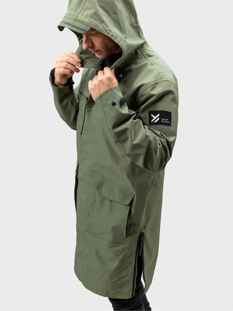 Man in Halti Tokoi II DX Parka, olive green, adjusting hood; black sleeve patch, plain backdrop.
