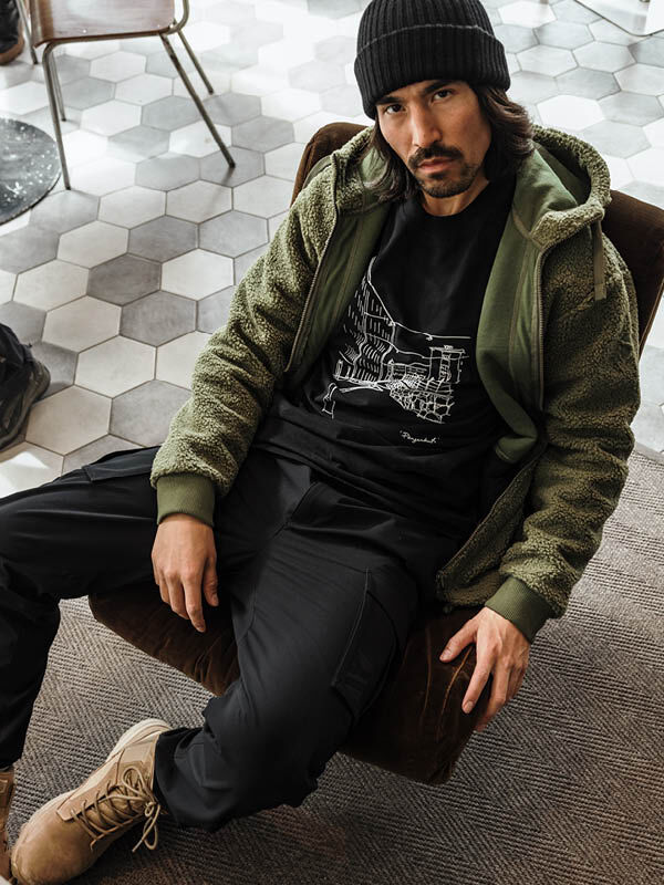 A man wears the Halti Lykky Fleece Jacket Mens, black pants, and boots while seated on a patterned tile floor.