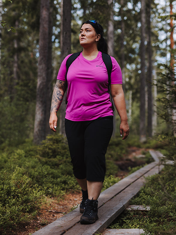 A woman in Halti Drive X-stretch Capri Pants hikes through a forest trail with a backpack.