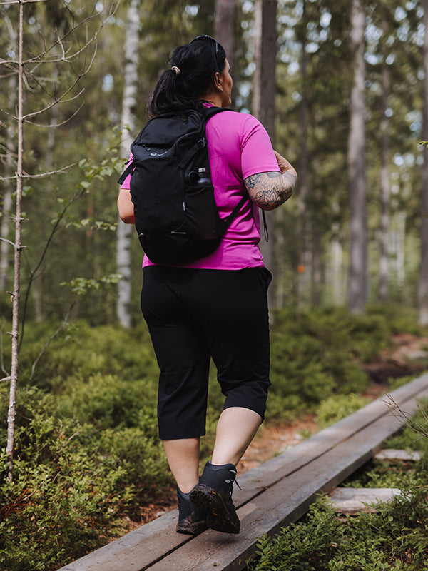 A woman in a pink shirt and black Halti Drive X-stretch Capri Pants walks on a wooden forest path with a black backpack.