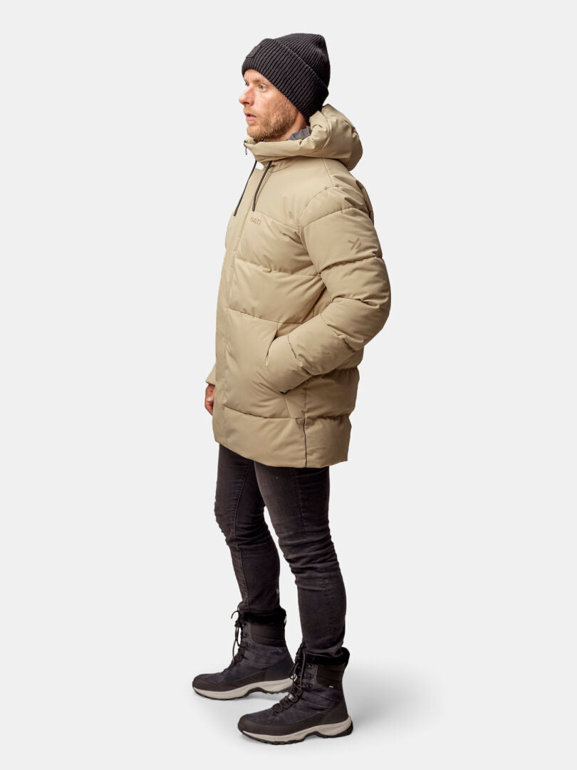 A man in profile wears the Halti Sammal Insulated Parka Jacket Men’s, black pants, boots, and a beanie on a white background.