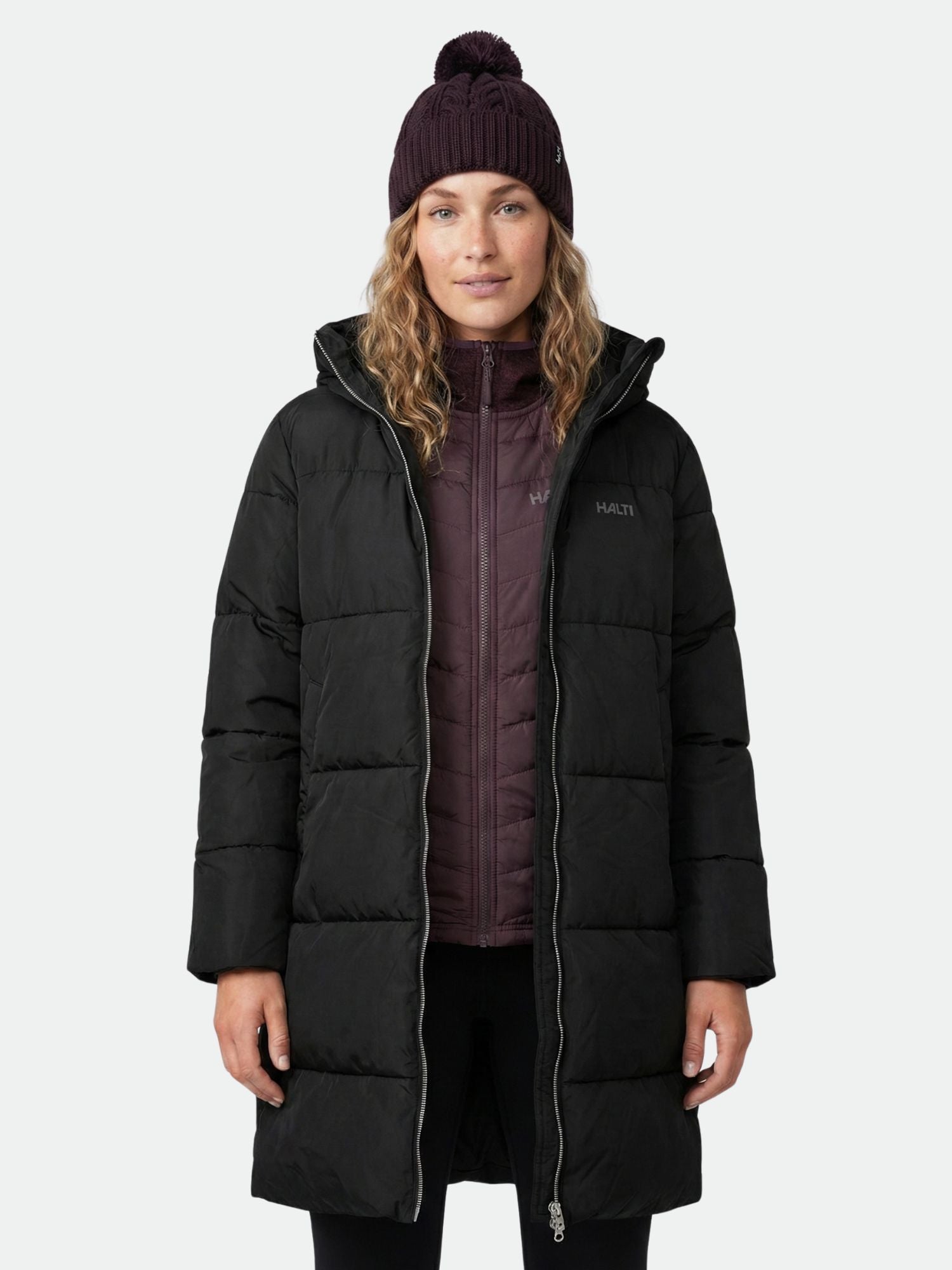 A woman wearing the Halti Sammal Insulated Parka Jacket in black, with a maroon jacket and knit beanie, facing forward.