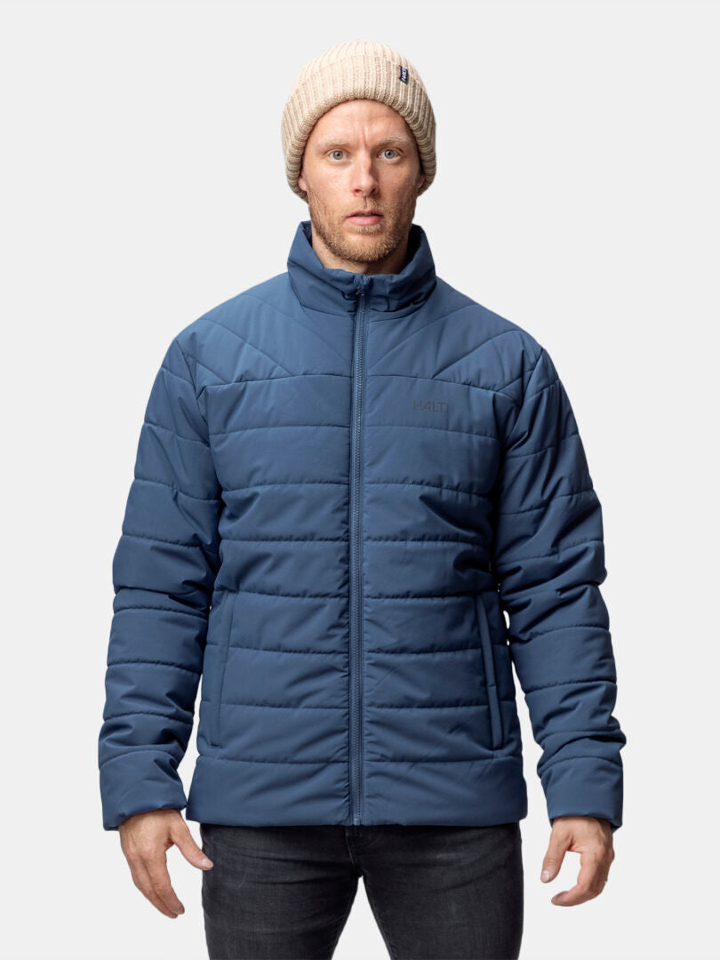 Man in a Halti Haapa Insulated Jacket, beige knit beanie, and dark jeans stands against a plain light background.