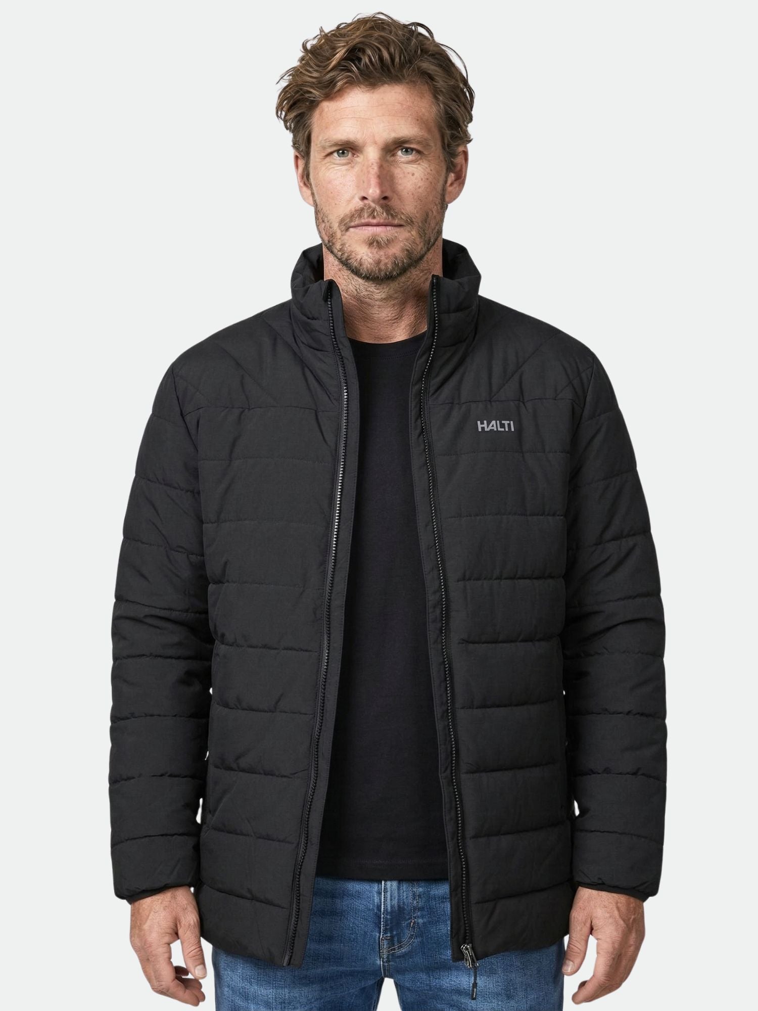 A man in a black Halti Haapa Insulated Jacket and blue jeans stands facing the camera against a plain background.
