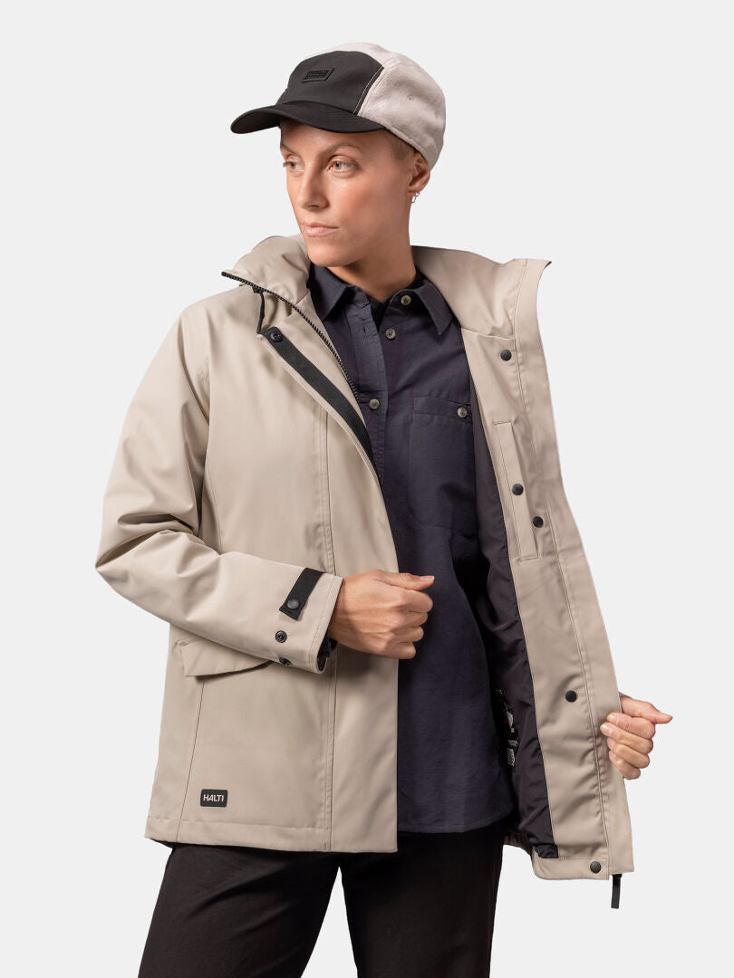A woman wears the Halti Tokoi DX Jacket in beige with a black shirt and two-tone cap against a plain light background.