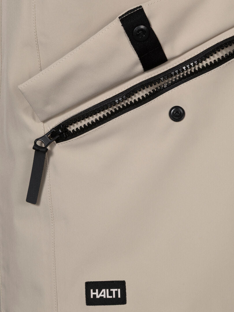 Close-up of Halti Tokoi DX Jacket Men's in beige DrymaxX 2-layer fabric with zip pocket, black Halti label, and buttoned strap.
