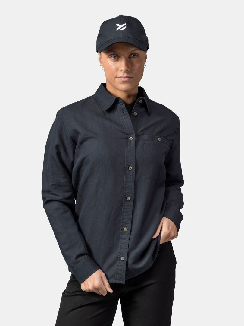 Woman wearing a Halti Notsgi Longsleeve Shirt, black pants, and a cap with white logo, standing against a plain background.