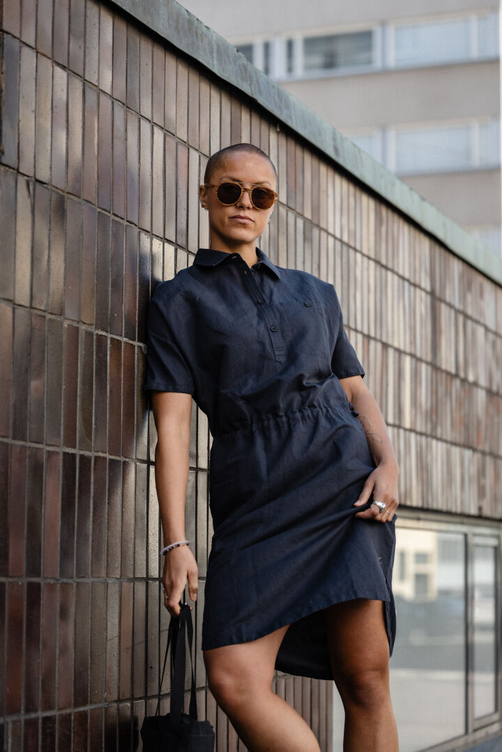 Woman in Halti Notsgi Dress and sunglasses stands by a tiled wall, holding a black bag.