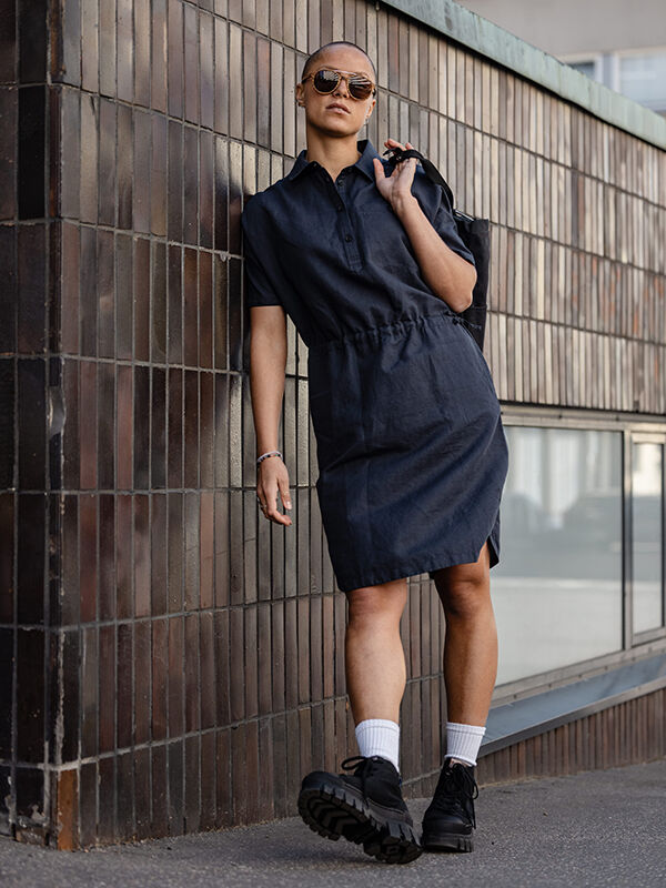 A woman in a Halti Notsgi Dress, sunglasses, and black boots leans against a tiled wall on a city sidewalk.