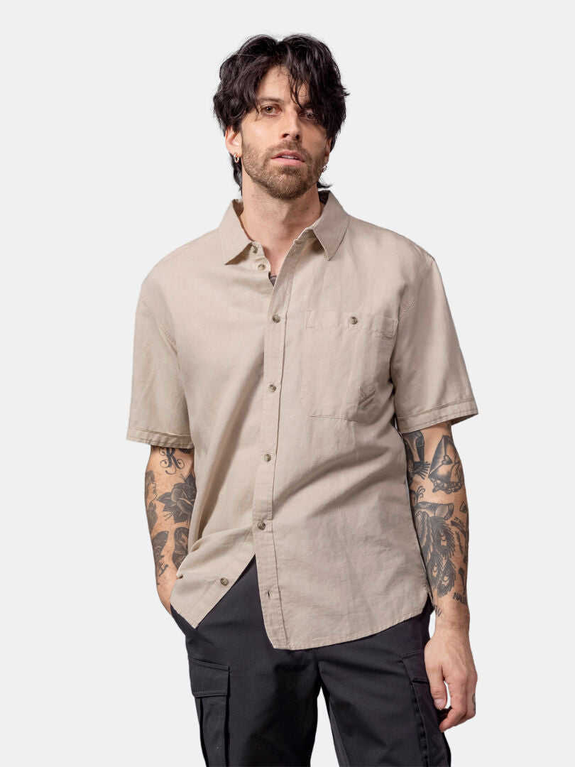 Man with dark hair and tattoos wears a beige Halti Notsgi Shirt Men's and black pants against a plain background.