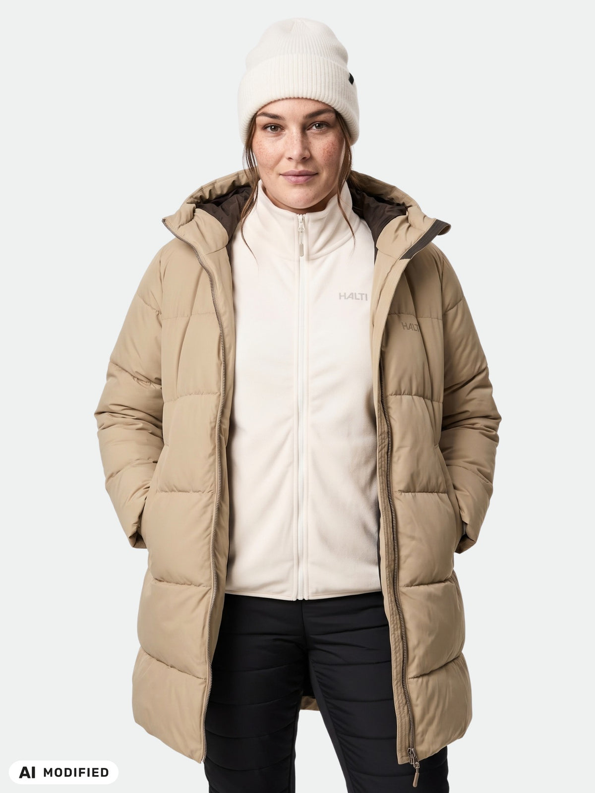 Woman wears Halti Sammal Insulated Parka Jacket Plus, white zip-up top, black pants, and beanie, hands in pockets.