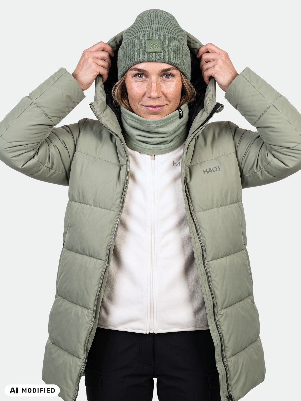 A woman in a green Halti Leinikko Puffer Parka, matching beanie, and neck warmer holds the hood with both hands.