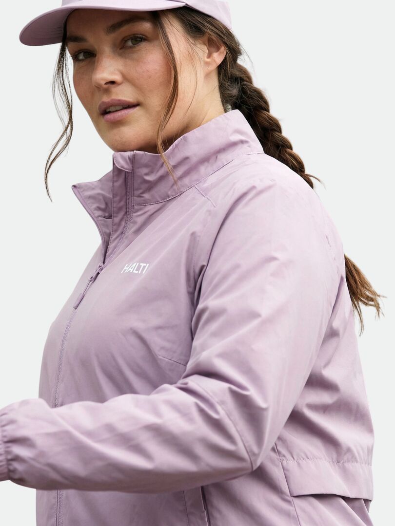 Woman in Halti's Saime Windbreaker Jacket Women's Plus and cap, with braided hair, stands against a neutral background.
