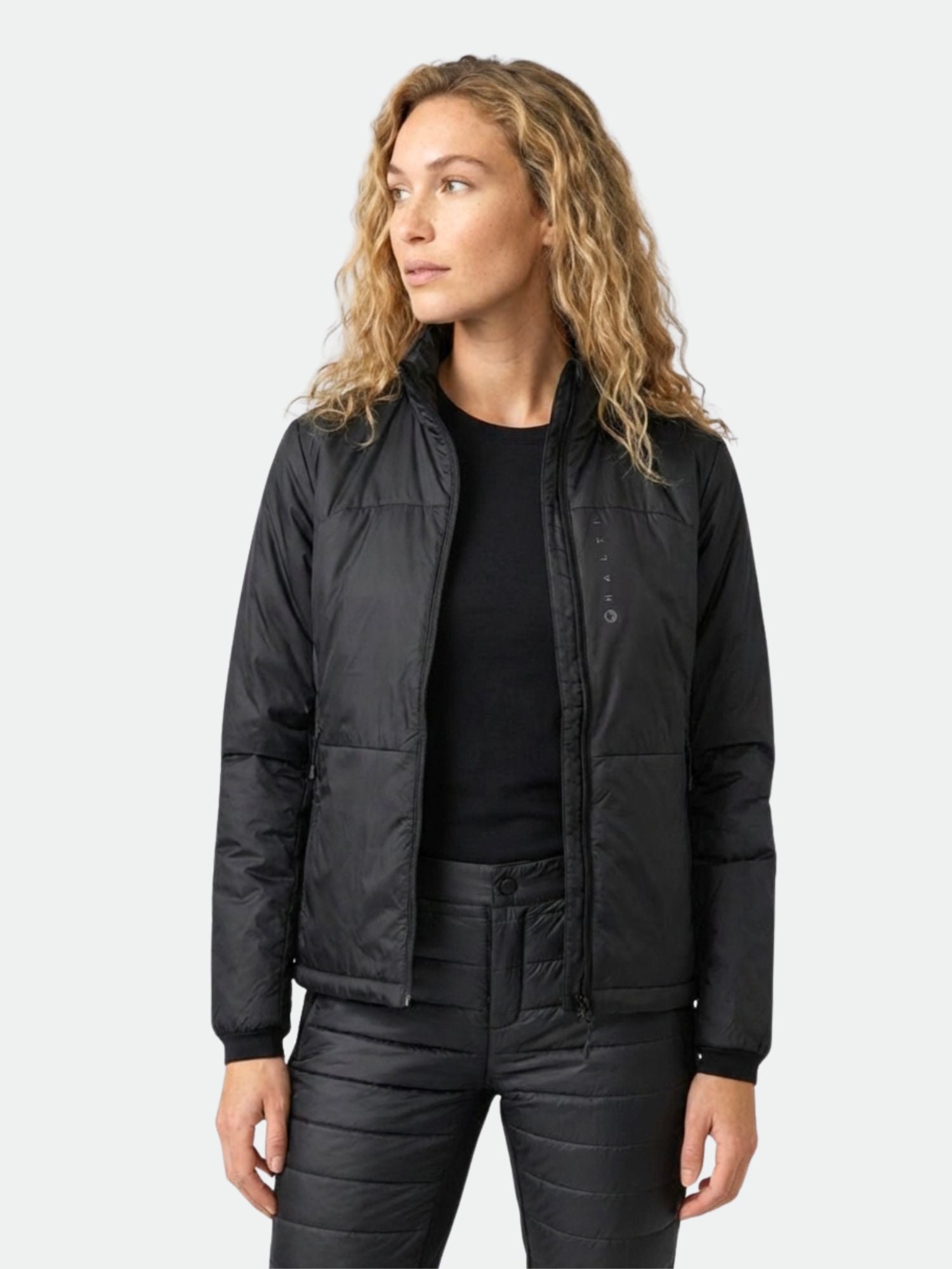 A woman with curly blonde hair wears a black Halti Neon Insulation Jacket, standing against a plain light background.