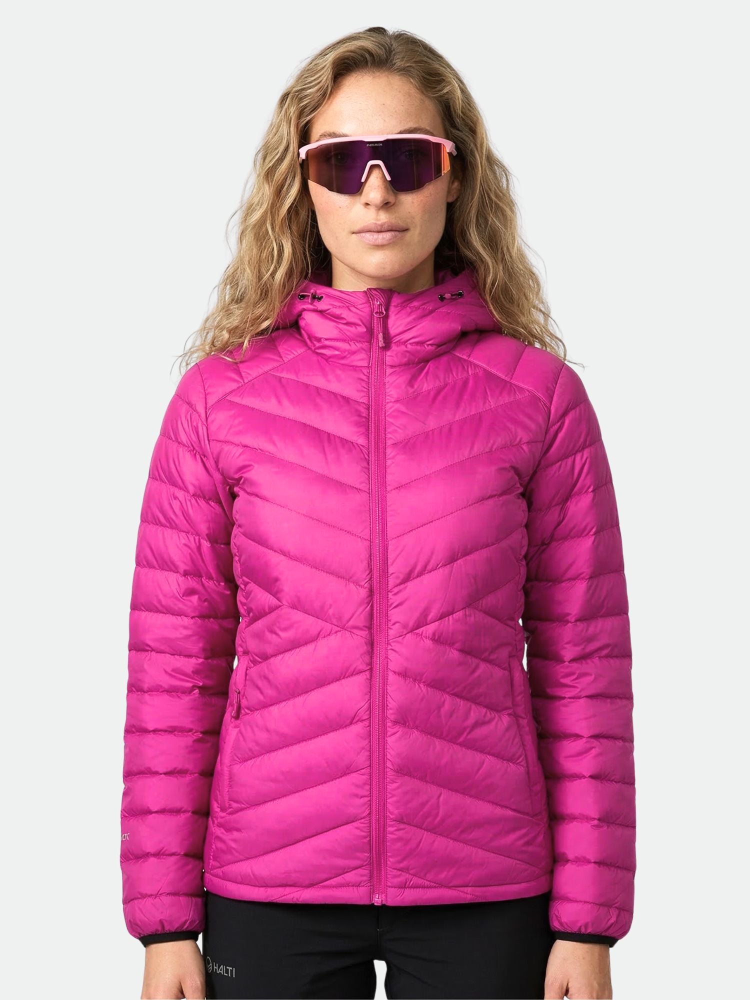 A woman wears the Halti Huippu Down Jacket in bright pink with large sunglasses, standing against a plain background.