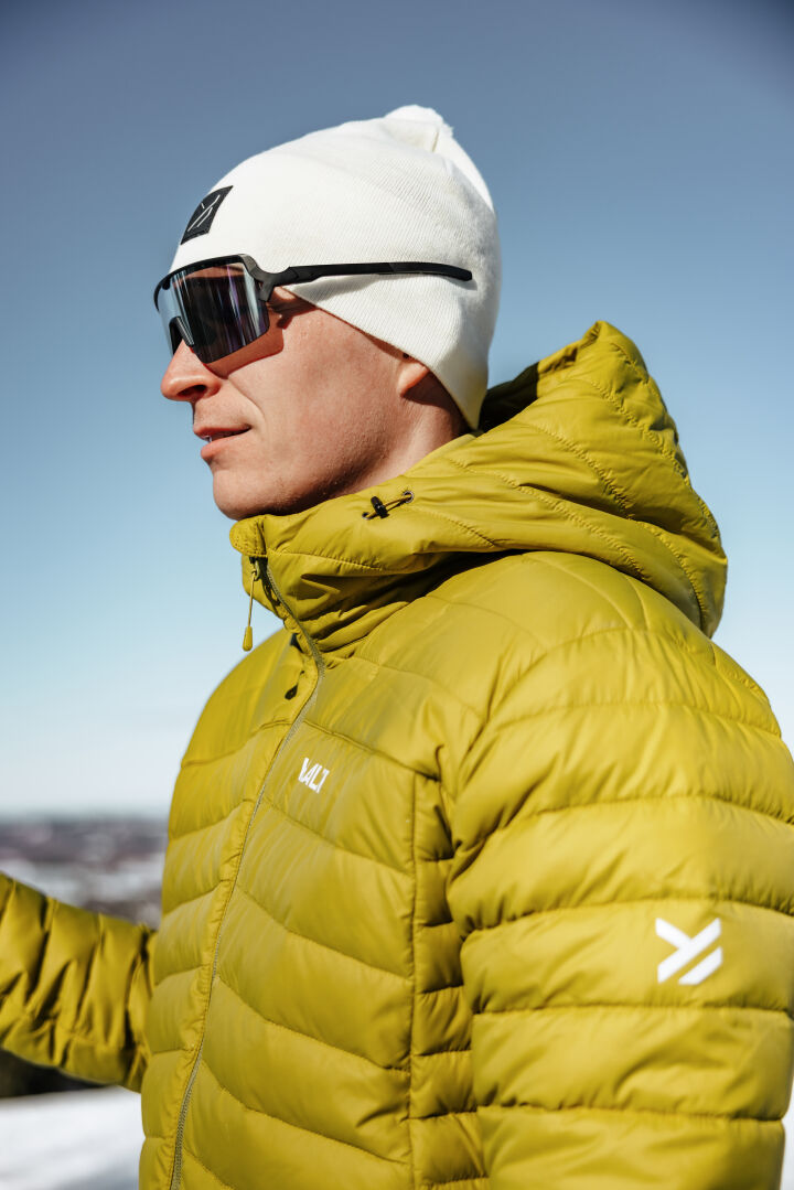 Person in a yellow puffer, Halti Pilke Beanie, and sunglasses stands outdoors on a sunny day.