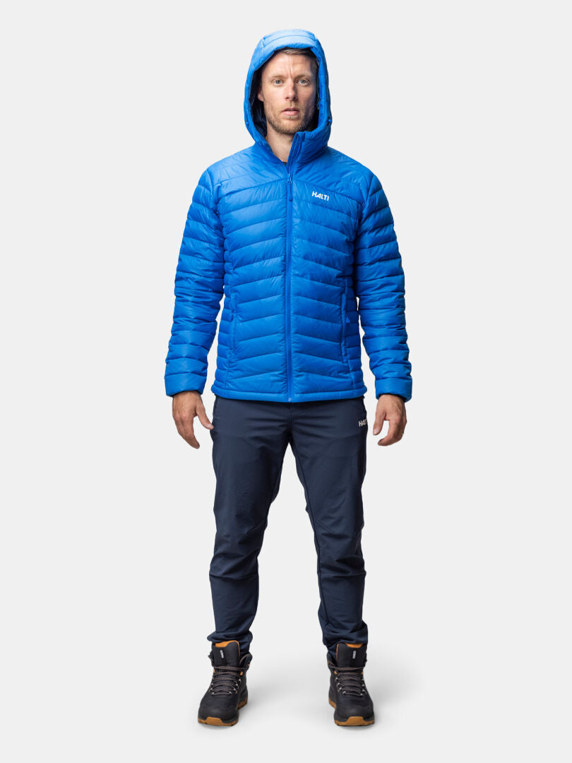 A man wears the Halti Huippu Down Jacket Men’s with dark pants and hiking boots against a plain background.