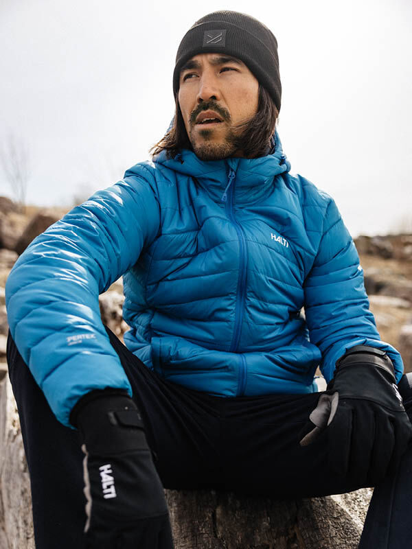 A man in a Halti Huippu Down Jacket and black beanie sits outdoors on a log, looking off to the side.