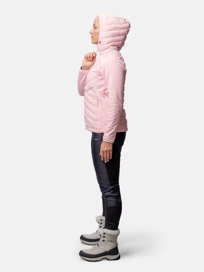 A woman in a pink Halti Huippu Hybrid Down Jacket, dark pants, and gray winter boots stands in profile.