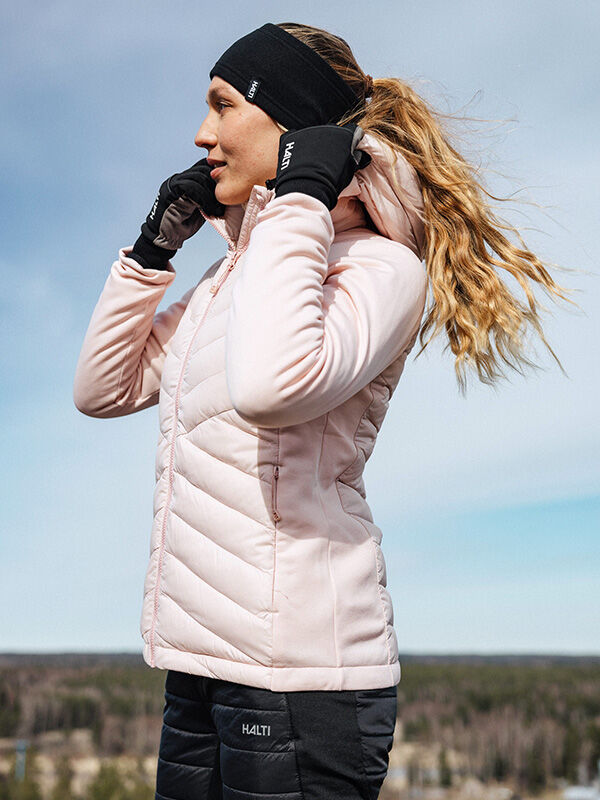 Woman wears Halti Huippu Hybrid Down Jacket in light pink, black gloves, and headband outdoors with trees and blue sky.