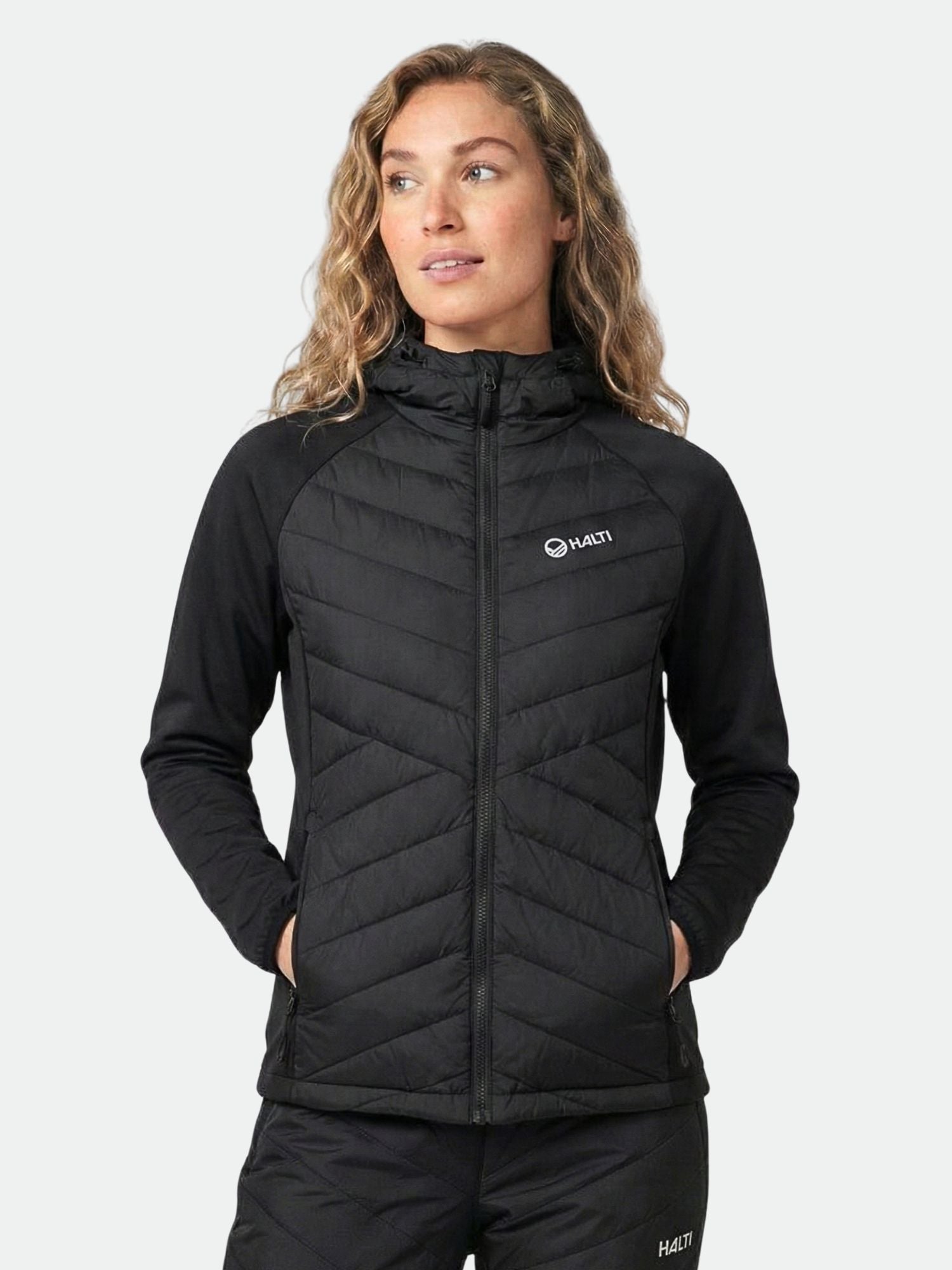 A woman wears the Halti Huippu Hybrid Down Jacket with hood, hands in pockets, looking slightly to the side.