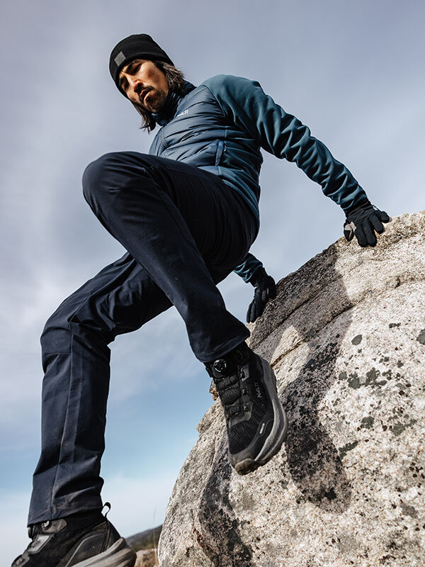 Wearing Halti Finholma X-stretch Pants, a man climbs over a large rock under a cloudy sky.