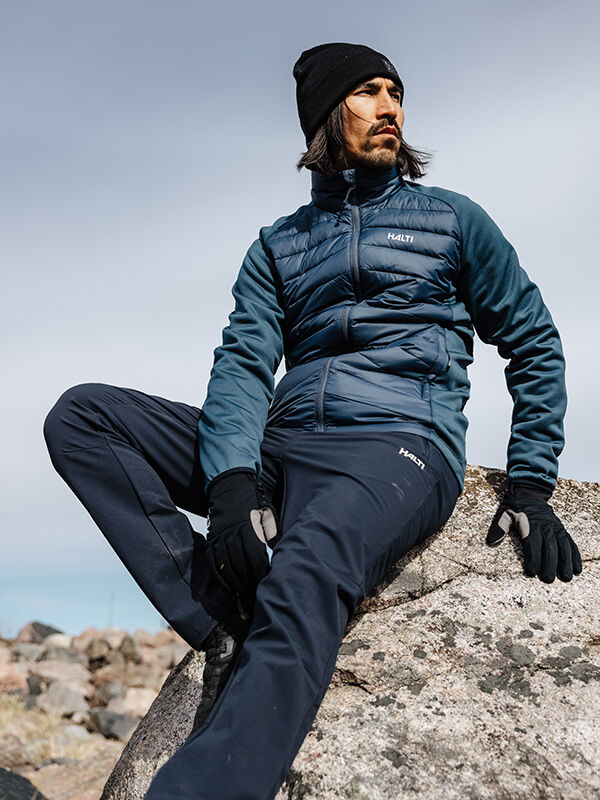A man in a Halti Huippu Hybrid Down Jacket, black hat, and gloves sits on a rock outdoors under cloudy skies.