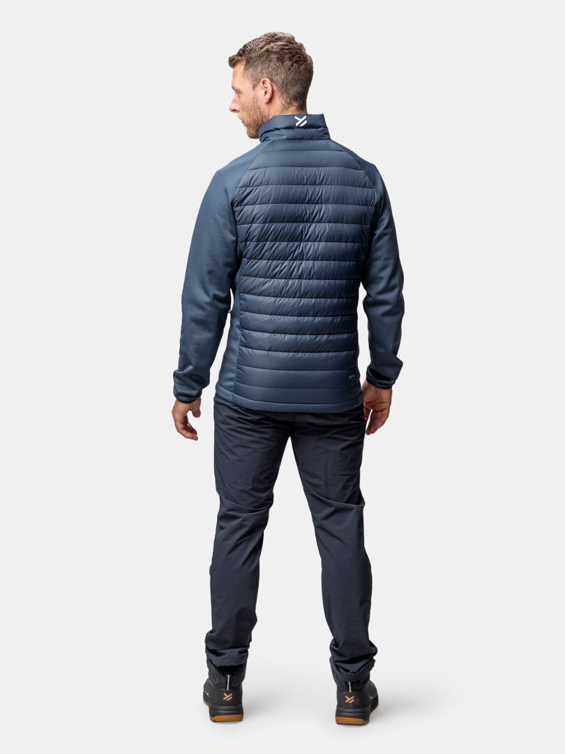 A man wears the Halti Huippu Hybrid Down Jacket (Mens), dark pants, and black shoes against a plain background.