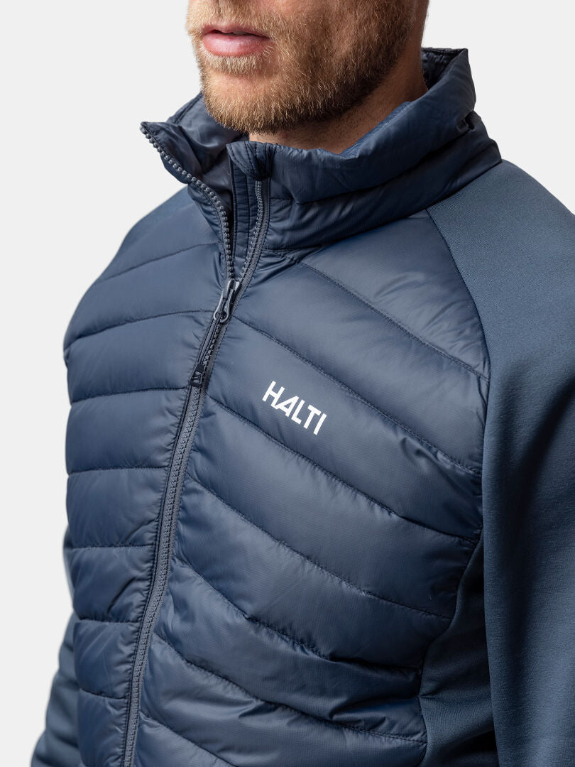 A man wears a Halti Huippu Hybrid Down Jacket with front zipper, high collar, and quilted design on a plain background.