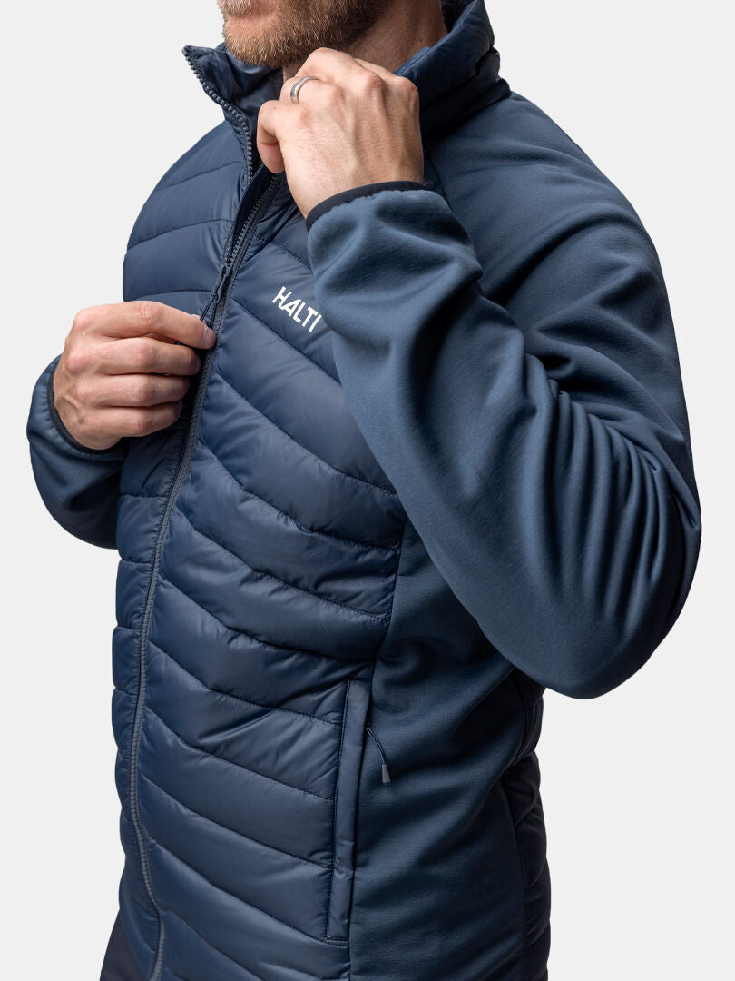 A man zips up a blue Halti Huippu Hybrid Down Jacket against a plain background.