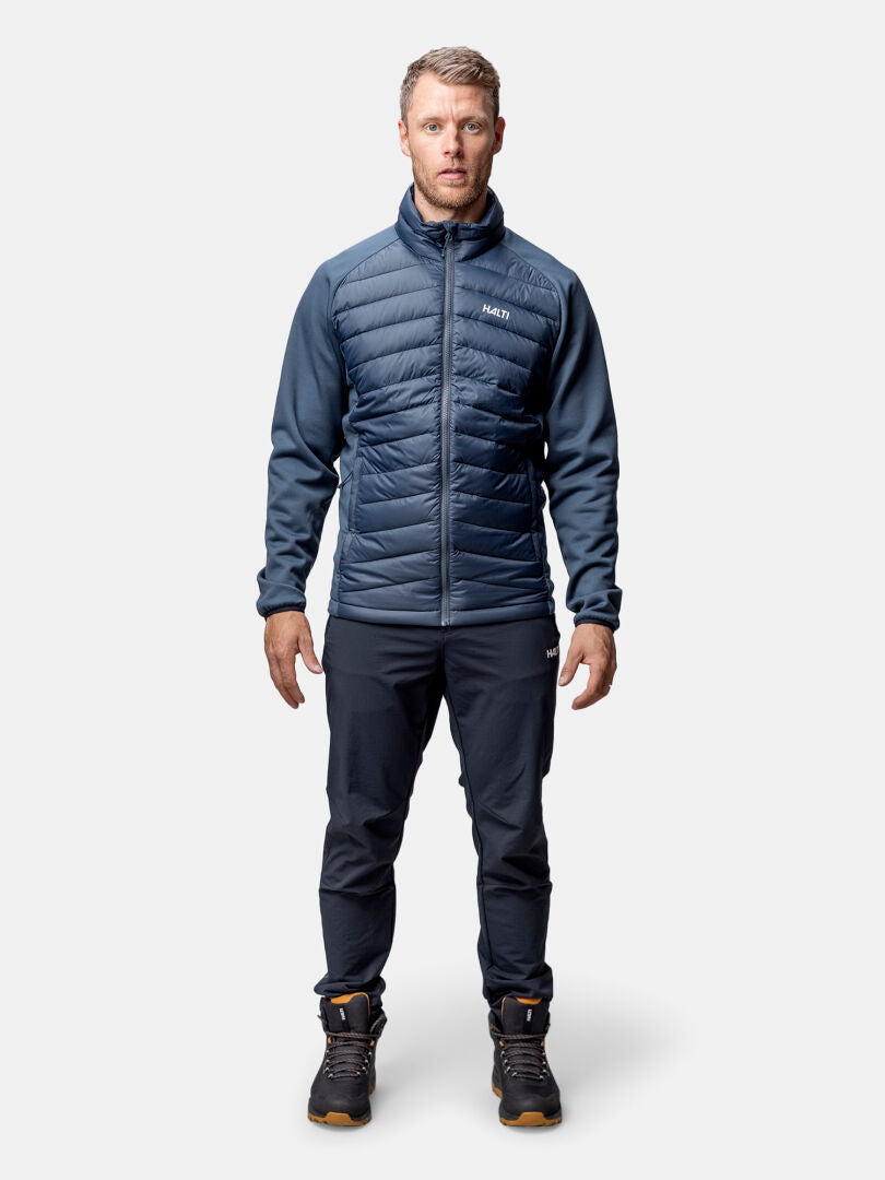 A man wears the Halti Huippu Hybrid Down Jacket Mens in navy, dark pants, and hiking boots against a plain background.
