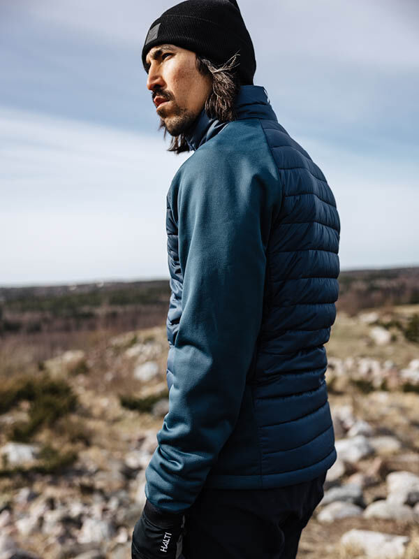 A man in a Halti Huippu Hybrid Down Jacket and black beanie stands on a rocky landscape, looking to the side.