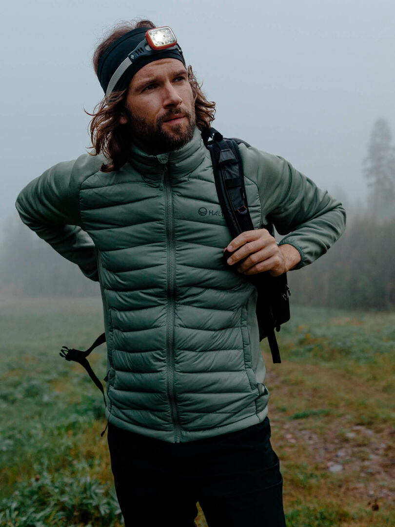 A man wearing the Halti Huippu Hybrid Down Jacket and a headlamp stands on a foggy trail with a backpack.