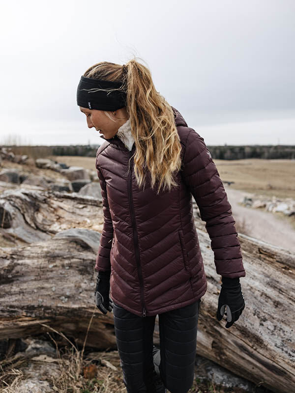Woman in a Halti Huippu Mid Down Jacket stands outdoors by a fallen tree, looking to her left.
