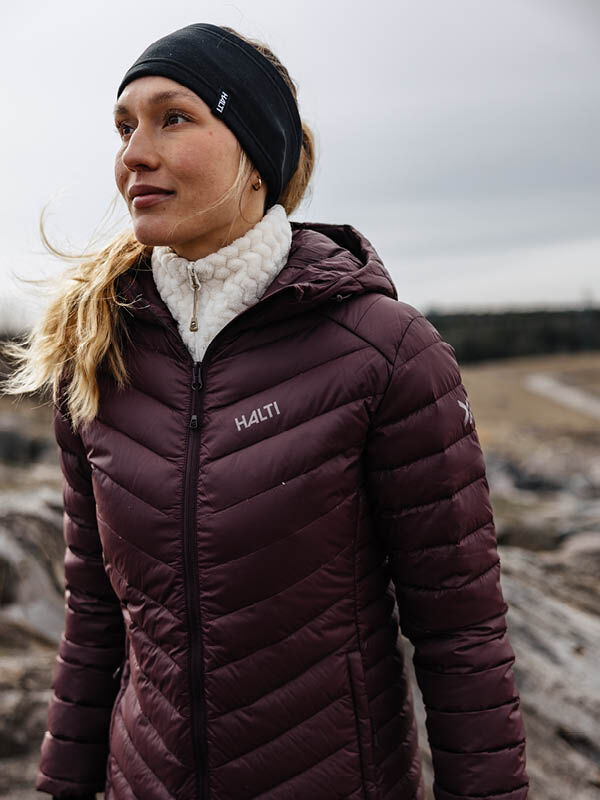 A woman outdoors wears a maroon Halti Huippu Mid Down Jacket, white sweater, and black headband, looking off to the side.