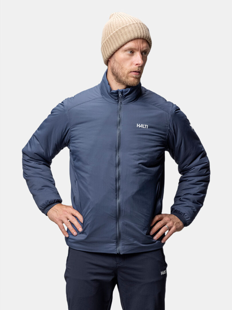 A man in a blue Halti Haave Insulation Primaloft Jacket stands hands on hips, looking left.