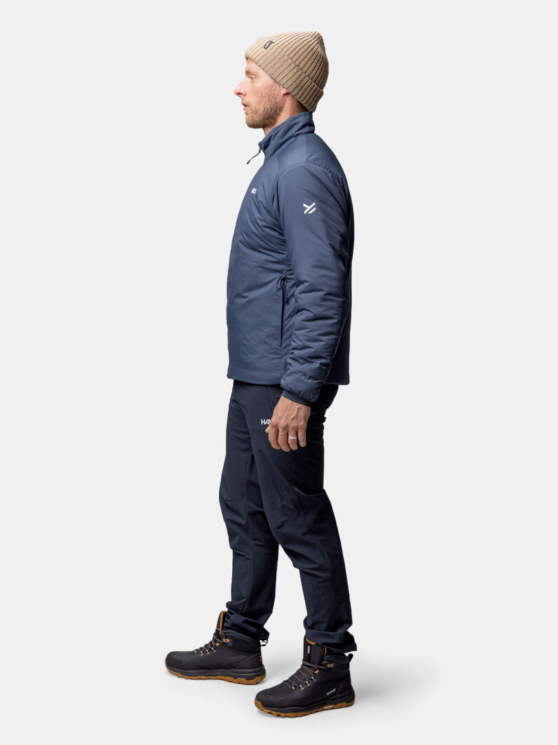 A man stands in profile wearing the Halti Haave Insulation Primaloft Jacket, blue pants, black boots, and a beige beanie.