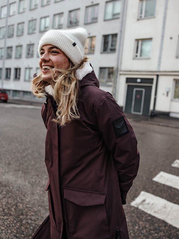 A person in a maroon coat and Halti Vuolle Mohair Beanie smiles outdoors with buildings in the background.