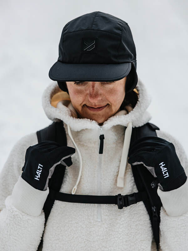 Person wearing a Halti Skutta Cap, white fleece jacket, and HALTI gloves, standing outdoors in the snow.