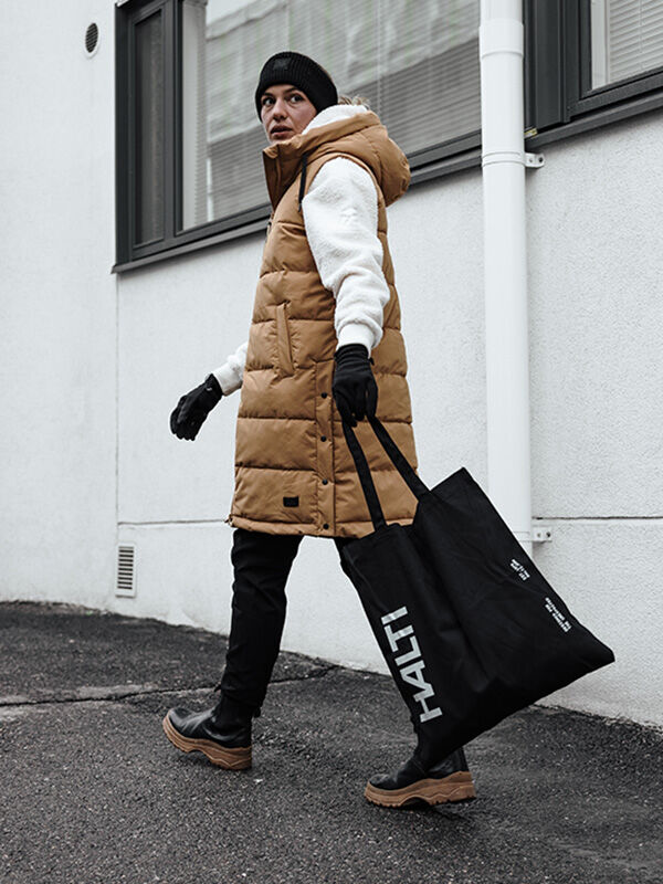 A person in a tan longline puffer vest, black Viiri Fleece Gloves, and a black beanie carries a large black tote bag.