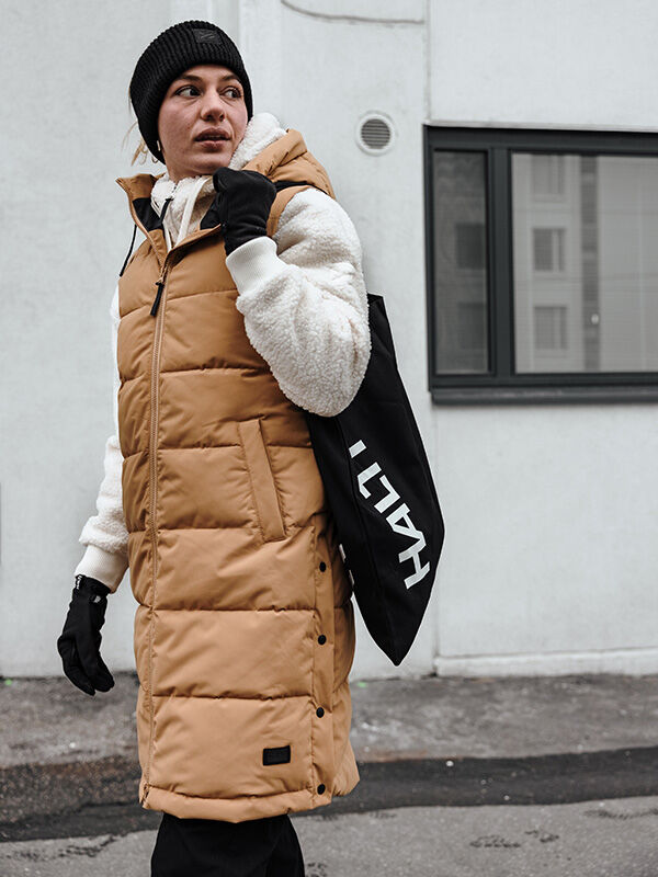A person in a tan puffer vest, white hoodie, and black beanie stands outdoors wearing Halti Viiri Fleece Gloves.