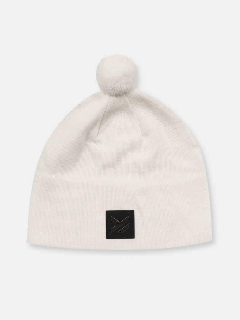 The Halti Pilke Beanie is a white merino wool hat with pom-pom and black patch, ideal for winter sports.