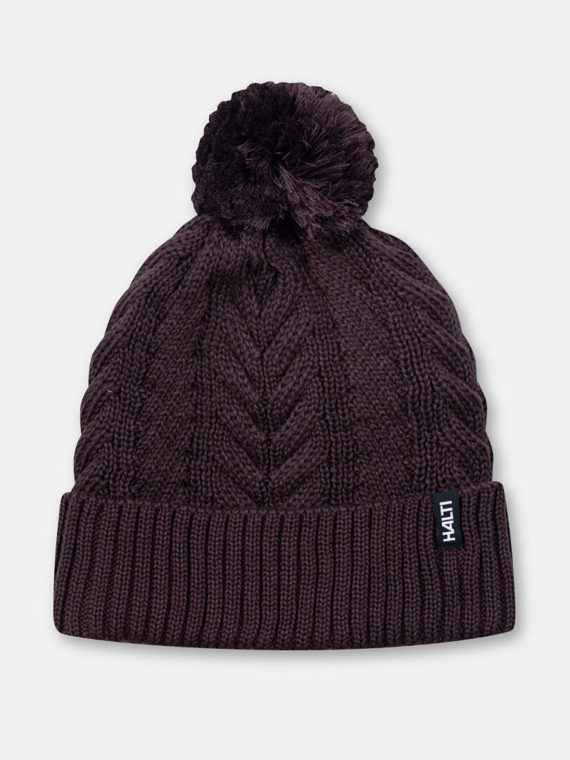 The Halti Hile Beanie is a dark brown cable knit hat with a pom-pom and “Haiti” tag—perfect for winter.