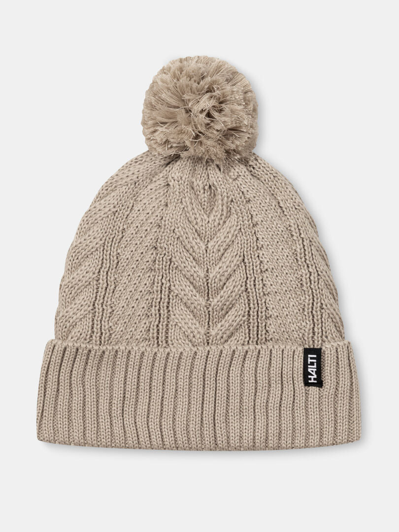 The Halti Hile Beanie is a beige cable knit hat with a pom-pom, folded brim, and black and white label.