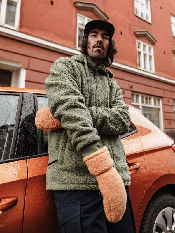 A man in Halti Lykky Fleece Mittens and a green fleece jacket stands by an orange car near a red building.