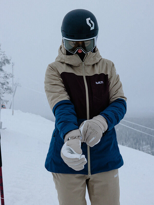 Wearing a ski jacket, helmet, and goggles, a person adjusts their Halti Vuotos Ski Gloves on a snowy slope.