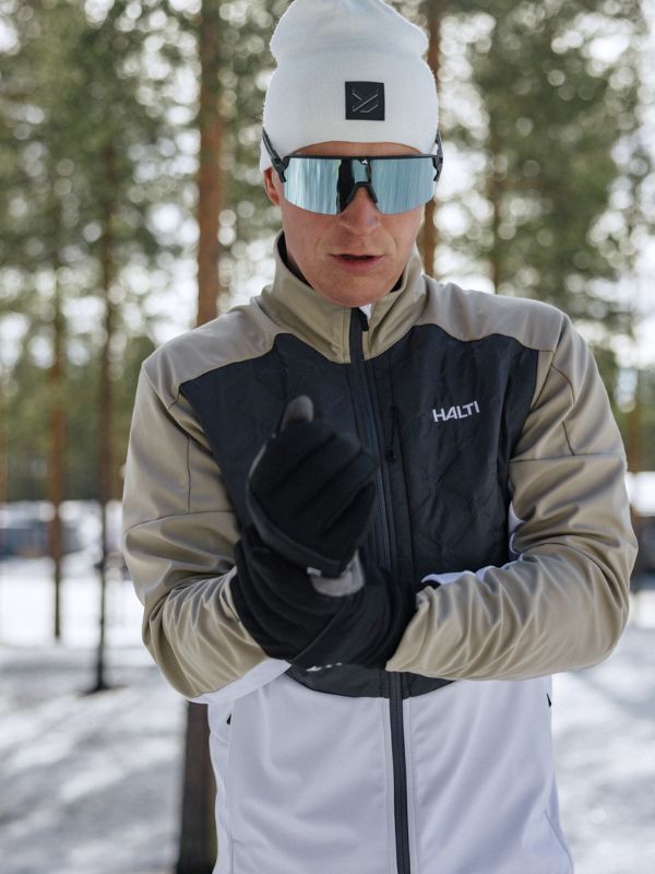 A man in a white beanie, sunglasses, and Halti jacket stands outdoors in snow, wearing Halti XC Touring II Gloves.