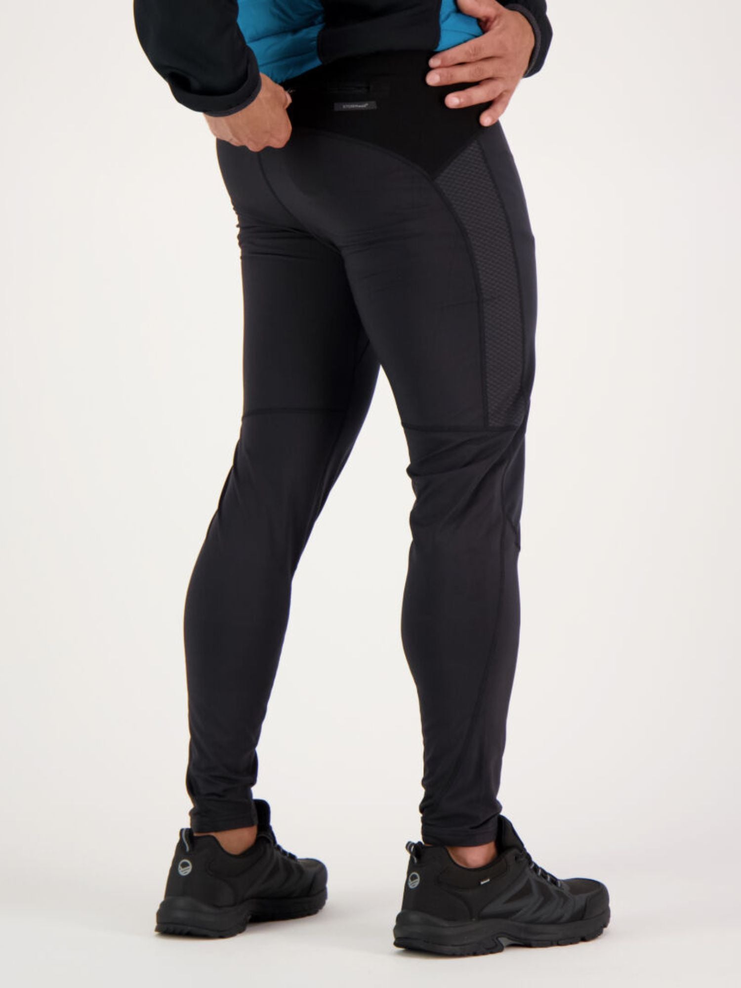 A person wears Halti Suunta Men's Hybrid Tights and black shoes, posing with one hand on hip against a plain background.