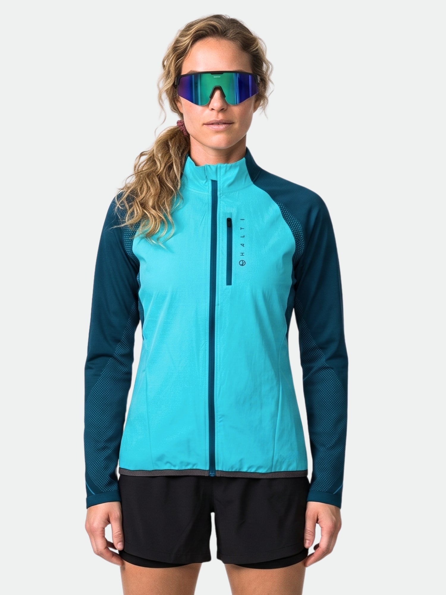 A woman wears the Halti Tracks Women's Seamless Jacket, black shorts, and reflective sunglasses against a plain background.